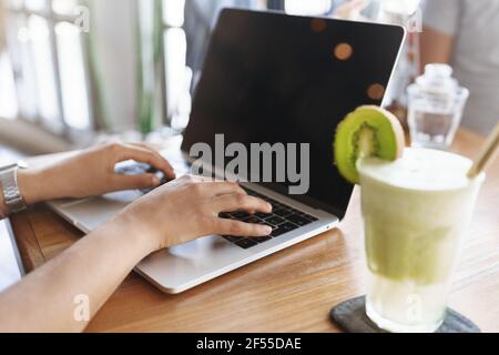 Cropped shot Frau Hände eingeben Laptop-Tastatur leeren Bildschirm, sitzen drinnen Café Tisch trinken grün Smoothie Cocktail Vorbereitung Universität Prüfung arbeiten Stockfoto