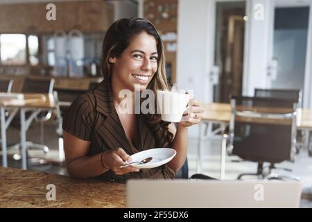 Fröhlich attraktiv gebräunt hispanische Frau sitzen Café kichern Spaß haben Genießen Sie angenehme unbeschwerte Gespräch Look Kamera liebenswert Lächeln halten klein Platte Stockfoto
