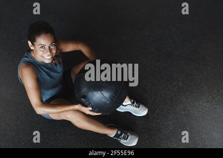 High-Angle-Ansicht glücklich motivierte brasilianische Frau Training, schauen Sie sich lächelnd halten Medizinball, Ruhe Fitness-Studio Boden nach produktiven Trainingseinheit Stockfoto