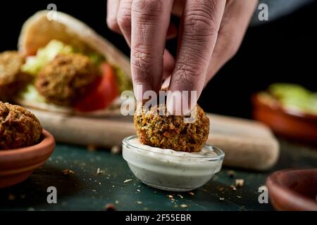 Ein junger Mann taucht einen Falafel in eine Schüssel Joghurtsauce an einem rustikalen grünen Holztisch Stockfoto