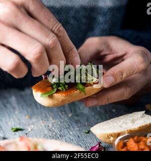 Ein junger Kaukasianer toppt mit gehacktem Salat eine vegane Vorspeise, die mit einem halbierten getoasteten Brotbrötchen mit einer Tomaten- und Mandelaufstrich bedeckt ist, bei einem Stockfoto