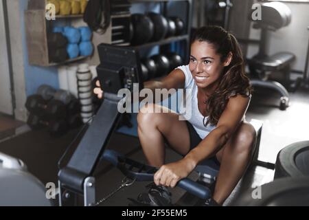 Side-Shot attraktive motivierte junge hispanic Sportlerin mit Beinpresse, Training auf funktionalen Trainingsgeräten, Pick-Programm auf dem Bildschirm, lächelnd Stockfoto