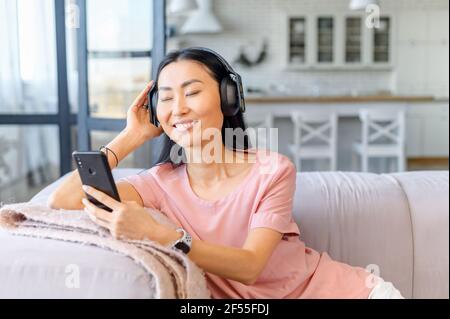 Eine entspannte asiatische Frau genießt Lieblingstitel zu Hause, eine Frau trägt Kopfhörer hält Smartphone und Tagträumen mit geschlossenen Augen Stockfoto