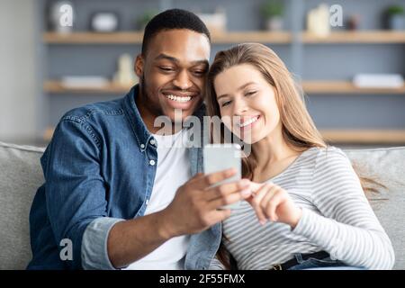 Fröhliches gemischtes Paar, das zu Hause Zeit mit dem Smartphone verbringt, Videos ansieht Stockfoto