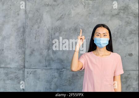 Eine asiatische Frau trägt schützende medizinische Maske zeigt Finger auf einen leeren Raum, neue soziale Regeln während Coronavirus-Pandemie, Studio-Foto isoliert auf grau, Kopierraum Stockfoto