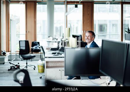 Lächelnder Geschäftsmann sitzt inmitten von Computern im Büro Stockfoto