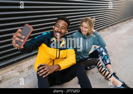 Fröhlicher Sportler mit Sportlerin, die während des Selfies über das Mobiltelefon unterwegs ist An der Wand sitzen Stockfoto