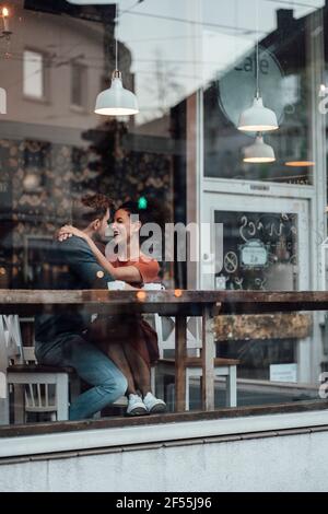 Lächelnde Frau, die den Mann ansieht, während sie mit dem Arm um sich herum sitzt Im Café Stockfoto