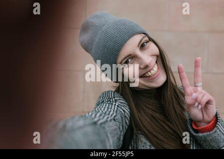 Fröhliche junge Frau in warmer Kleidung, gegen die ein Friedenszeichen angezeigt wird Wand Stockfoto