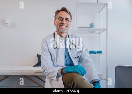 Hausarzt mit Schutzhandschuh lächelnd, während er in der Klinik sitzt Stockfoto