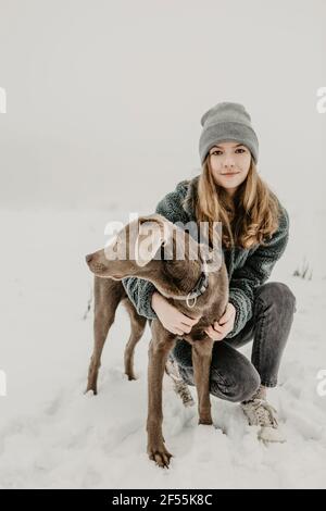 Porträt eines Teenagers, das im Schnee hockte und Labrador umarmte Abrufen Stockfoto