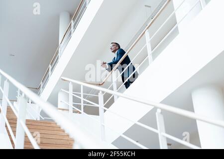 Ein leitender männlicher Geschäftsexperte blickt weg, während er sich an Geländer lehnt Im Flur im Büro Stockfoto