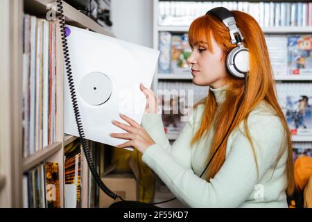 Rothaarige Frau mit Kopfhörern sucht nach Musik aus dem Regal Speichern Stockfoto