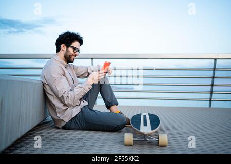 Lächelnder Geschäftsmann, der auf dem Longboard ein Mobiltelefon benutzt, während er sitzt pier lehnt sich an die Bank Stockfoto