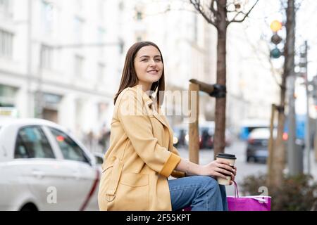 Lächelnde junge Frau hält Einweg-Kaffeetasse, während sie im Sitzen sitzt Stadt Stockfoto