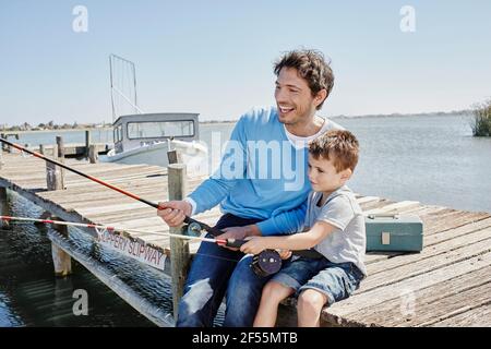 Fröhlicher Vater, der mit seinem Sohn angeln kann, während er auf dem Pier sitzt Stockfoto