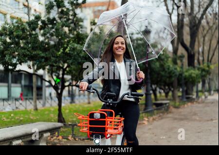 Schöne Frau, die auf einem elektrischen Fahrrad fährt, während sie einen Regenschirm auf der Straße in der Stadt hält Stockfoto