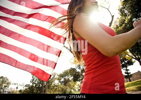 Junge Frau, die im Park läuft, während sie die amerikanische Flagge hält Sonniger Tag Stockfoto