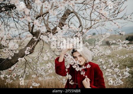 Blonde Frau berührt den Ast, während sie unter dem blühenden Mandelbaum steht Stockfoto