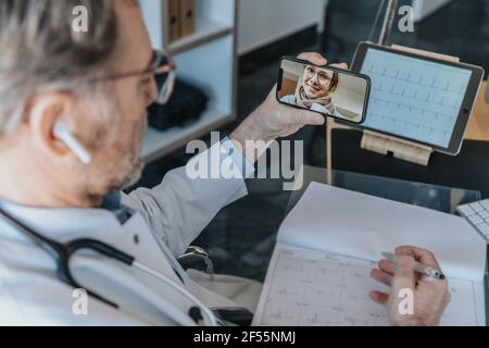 Reifer Arzt im Gespräch mit weiblichen Patienten über Mobiltelefon, während Sitzen in der Arztpraxis Stockfoto