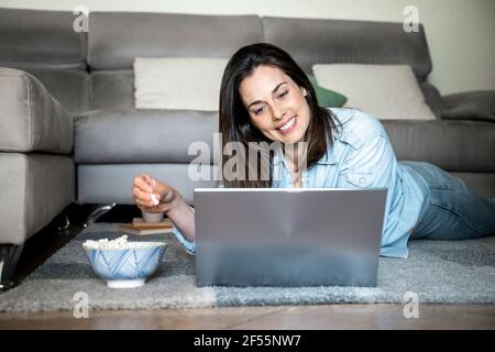 Lächelnde Frau mit Laptop, die Popcorn isst, während sie auf dem Teppich liegt Zu Hause Stockfoto