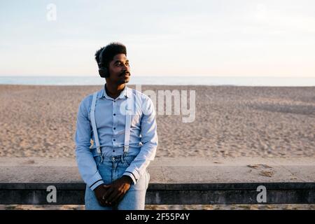 Mann lehnte sich am Strand an einer Stützmauer, während er Musik hörte Über Kopfhörer Stockfoto