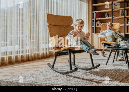 Lächelnder Vater, der den Sohn gestikuliert ansieht, während er auf dem Stuhl sitzt Im Wohnzimmer Stockfoto