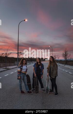 Lächelnde Freundinnen mit Skateboards, die während des Sonnenuntergangs auf der Straße stehen Stockfoto