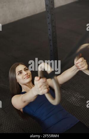 Vertikaler Oberwinkel-Schuss lächelnd, stark und energiegeladen, motivierte Frau Workout im Fitnessstudio, hängen an Gymnastikringen, Functional Training Workout Stockfoto