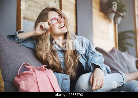 Ja, ich fühle mich wie die Königin dieser Stadt. Außenaufnahme einer gutaussehenden selbstbewussten blonden Frau in Denim, die auf dem Sofa sitzt und eine kühlende, berührende Sonnenbrille Stockfoto