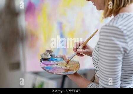 Weibliche Künstlerin, die Farben auf Palette mischt, während sie abstrakte Leinwand malt Zu Hause im Studio Stockfoto