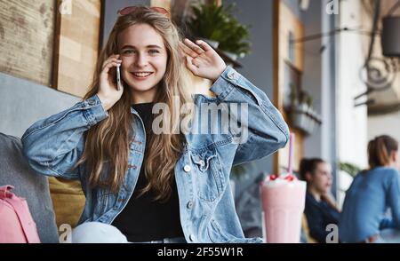 Junge Frau, die auf dem Handy spricht, während sie allein im Café sitzt, auf Freund verzichtet, Smoothie trinkt und auf der Restaurantterrasse ruht Stockfoto