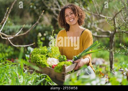 Lächelnde Frau in einer Kiste mit frisch gepflücktem Bio-Gemüse In Permakultur Garten Stockfoto