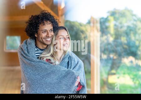Lächelndes Paar, das mit einer Decke bedeckt war, während es am Fenster zusammenstand Im Vorgarten Stockfoto