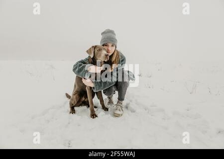 Porträt eines Teenagers, das im Schnee hockte und Labrador umarmte Abrufen Stockfoto