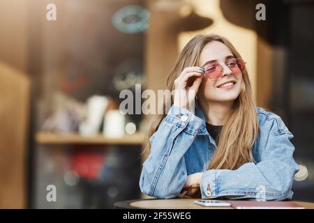 Stil liegt ihr im Blut. Selbstbewusst gut aussehende urbane Mädchen mit blonden Haaren in trendigen Sonnenbrillen und Denim-Jacke, lehnte sich auf dem Tisch während des Stehens und Stockfoto