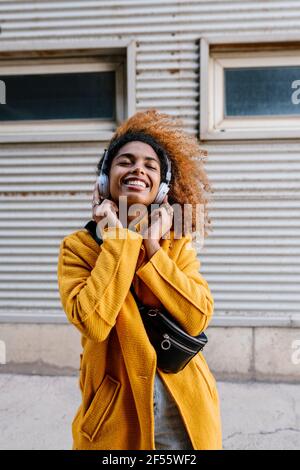 Lächelnde Afro-Frau mit geschlossenen Augen, die über Kopfhörer Musik hört Gegen die Wand Stockfoto