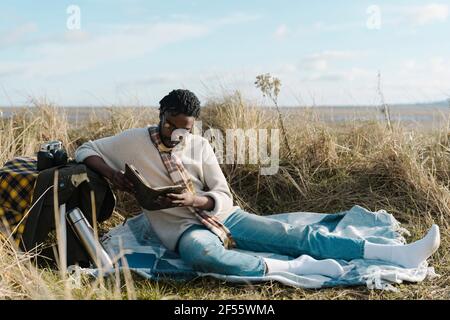 Junger Mann liest Buch, während er am Strand auf der Decke sitzt Es weht schwacher bewölktes Wetter Stockfoto