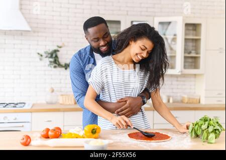 Liebevolles junges multirassisches schwarzes Paar, das Pizza zusammen an der Küchentheke macht, junger Mann, der mit Liebe seine zarte Frau umarmt, die den Teig mit Tomatenmark verteilt und Essen zusammen zu Hause kocht Stockfoto