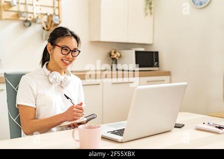 Junge Frau mit Kopfhörern, die während der Arbeit in Notizblock schreiben Auf dem Laptop zu Hause Stockfoto
