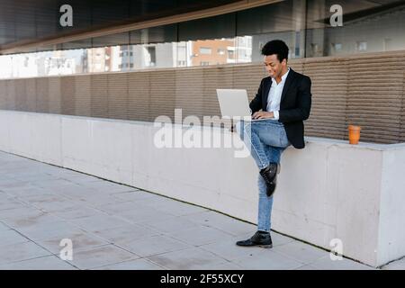 Männlicher Unternehmer mit Laptop, während er auf einem Bein gegen die Haltewand steht Stockfoto