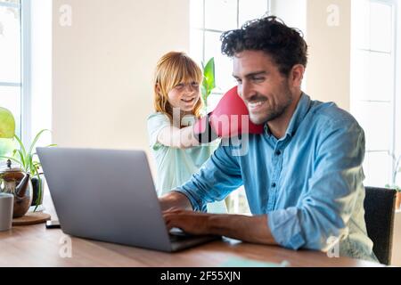 Lächelndes Mädchen mit rothaarige Mädchen mit Boxhandschuhen, die den Vater bei der Arbeit schlagen Laptop zu Hause Stockfoto