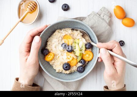 Die Hand des Weibchens schaufelt einen Haferbrei mit Heidelbeeren und Früchten. Gesundes Frühstück auf einem weißen Tisch. Stockfoto