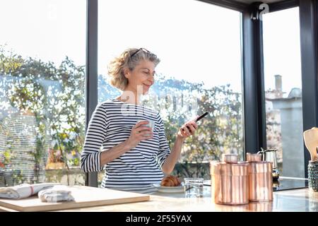 Lächelnde Frau mit Kaffeetasse und Mobiltelefon im Stehen Zu Hause Stockfoto