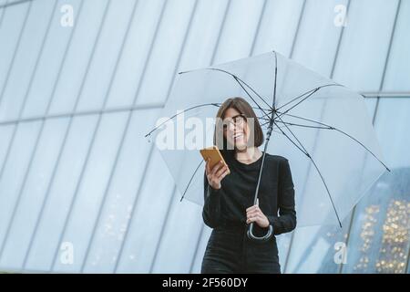 Fröhliche junge Frau mit Regenschirm und Smartphone im Stehen Vor dem Glasgebäude Stockfoto