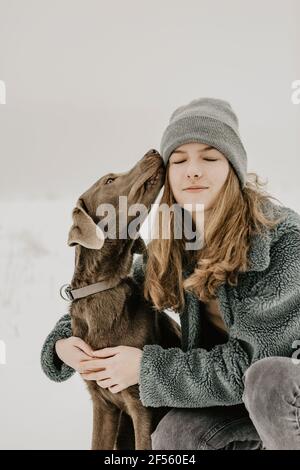 Porträt eines Teenagers, das im Schnee hockte und Labrador umarmte Abrufen Stockfoto