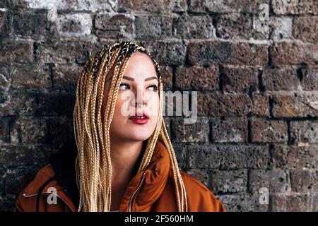 Frau mit langen geflochtenen Haaren schaut weg, während sie sich anlehnt Backsteinmauer Stockfoto