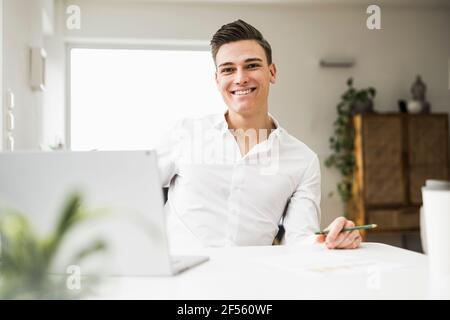 Lächelnder junger Mann mit Laptop am Schreibtisch Stockfoto