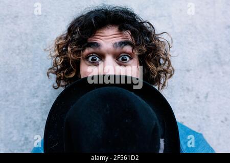 Junger Mann mit erhobenen Augenbrauen, der Hut vor sich hält Gesicht vor der Wand Stockfoto