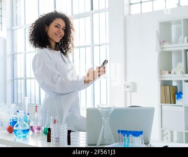 Lächelnde Wissenschaftlerin mit Smartphone, während sie sich auf den Tisch lehnte Im Krankenhaus Stockfoto